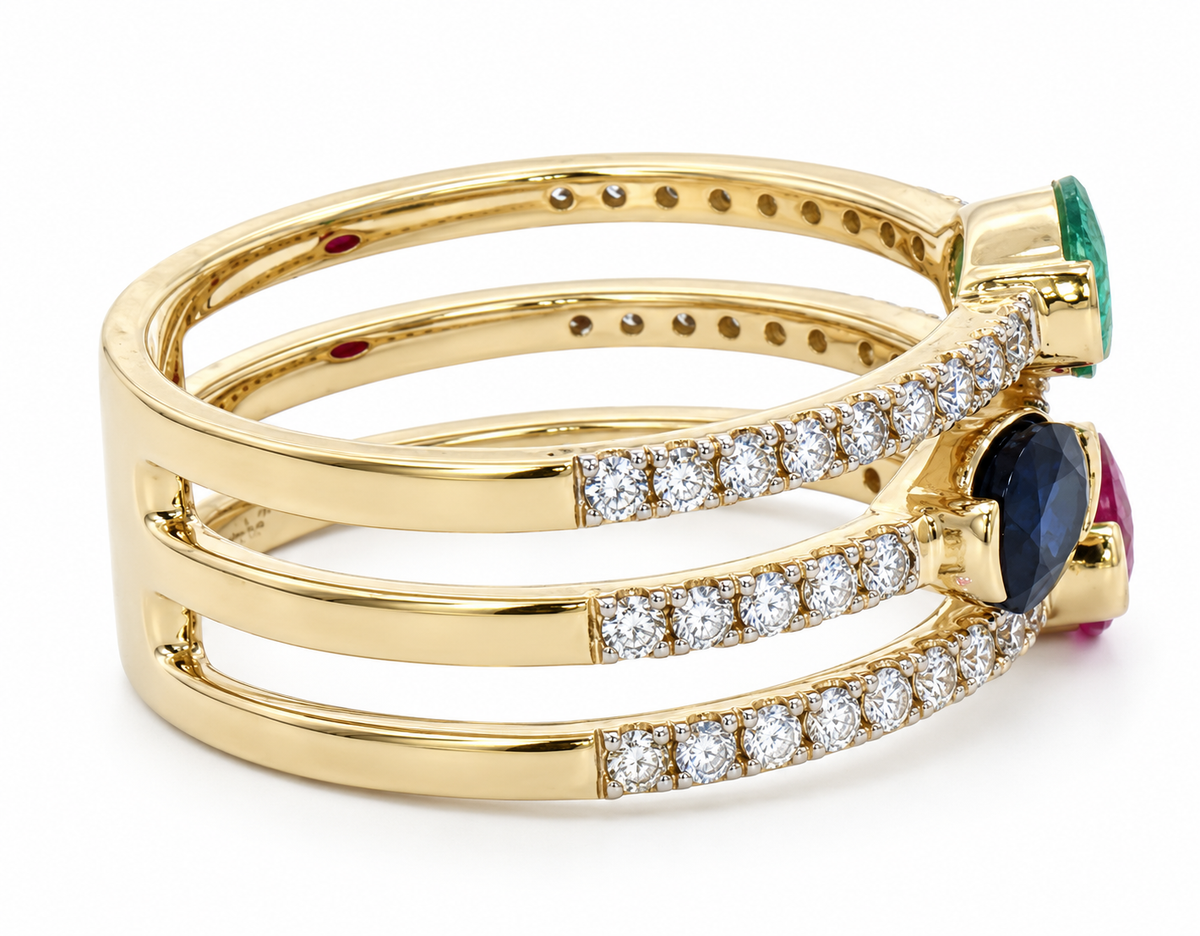 A gold triple-band ring featuring pavé-set round diamonds, a large oval-cut dark blue sapphire, a smaller oval-cut pink ruby, and a vertical green emerald-cut stone, shown on a white background.