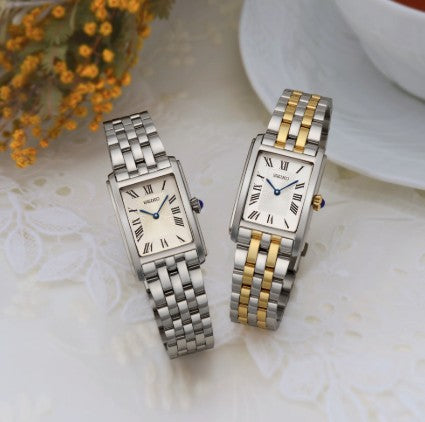 Two Seiko Essentials SS TT watches with stainless steel bracelets rest on lace, one all silver, one with gold accents.