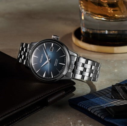 A Seiko Presage Cocktail Time SS Automatic Blue Dial Watch sits on a table beside a wallet and drink.