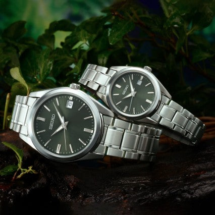 Two Seiko Essentials SS Quartz Green Dial Watches on a dark surface, surrounded by lush green foliage.
