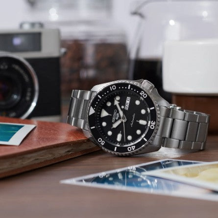 A Seiko 5 Sports SS Automatic Black Dial Watch by Seiko sits on a table with photos, a camera, and a cup behind.