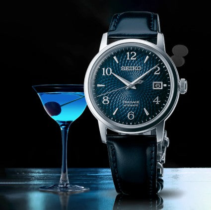 A Seiko Presage Cocktail Time Watch by Seiko with a blue dial is beside a blue cocktail with an olive on a glossy surface.