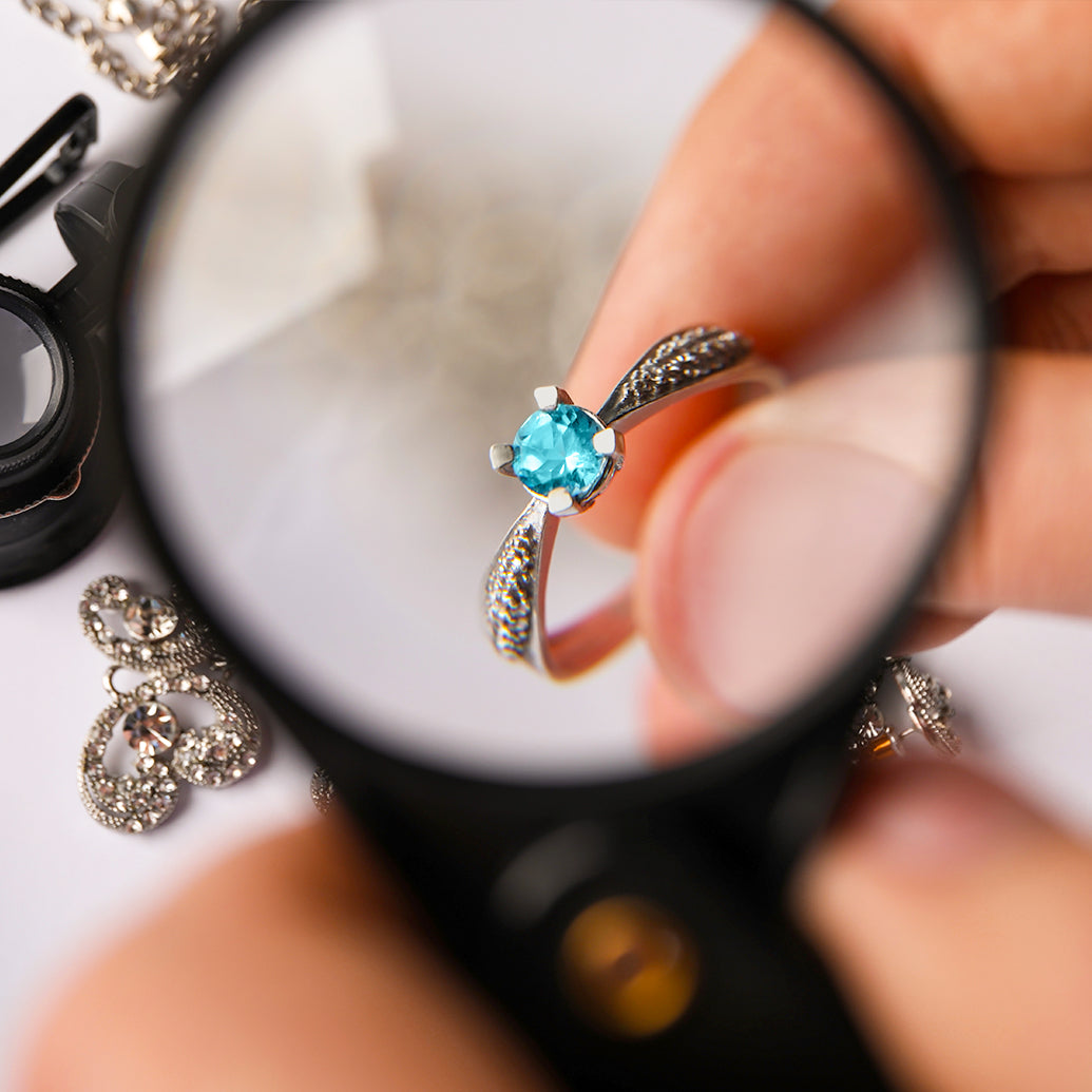 Close-up of a turquoise gemstone ring under a magnifying glass, with jewelry in the background.