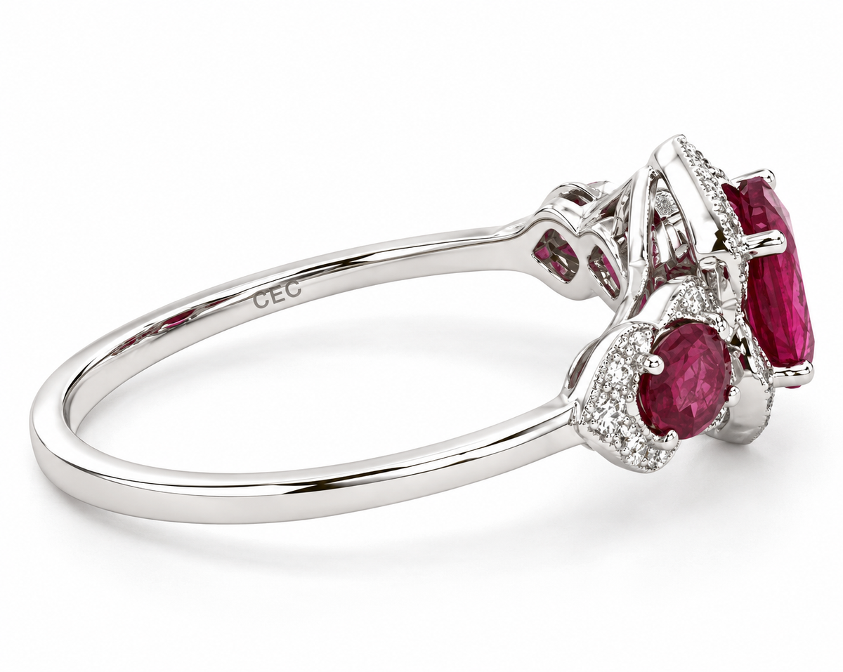 A white gold three-stone ring featuring a large oval-cut pinkish-red ruby center stone, flanked by two smaller oval ruby stones, all surrounded by small clear pavé diamonds.