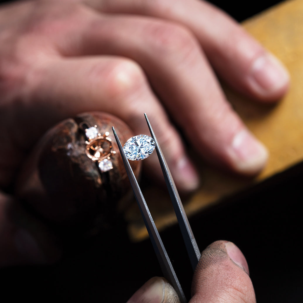 Close-up of a diamond being held with tweezers, with a blurred background of a hand holding the ring that the diamond will be placed on