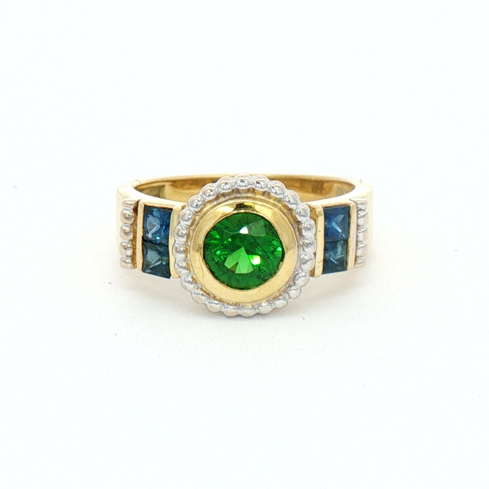 A yellow gold ring featuring a round green emerald center stone, surrounded by a halo of small clear stones, with blue rectangular side stones.