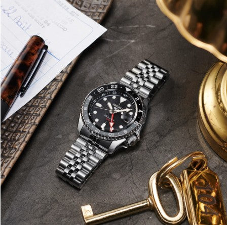 A Seiko 5 Sports SKX Sports Style GMT Series watch by Seiko sits on a desk with a pen, paper, and brass key.