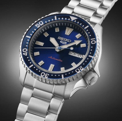 Seiko 5 Sports SS Automatic Blue Dial Watch with day-date and stainless steel bracelet, on a dark gradient background.