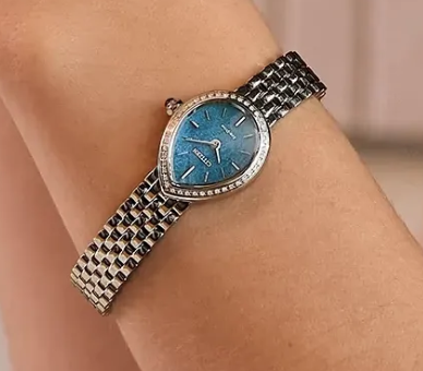 A close-up of a women's watch with a textured blue dial, a bezel set with small clear stones, and a silver-toned mesh bracelet, shown worn on a wrist.