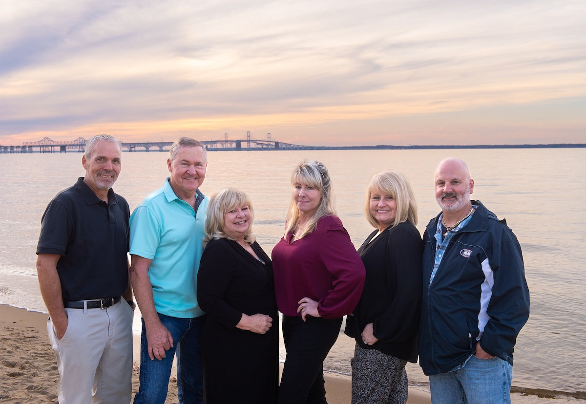 Six members of the Kent Island Jewelry team standing on a beach with a sunset and the Chesapeake Bay Bridge in the background