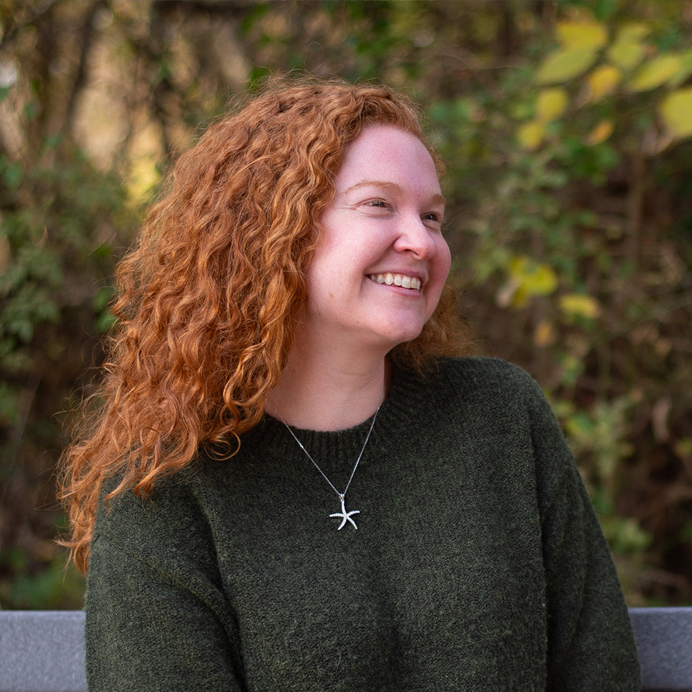 Woman with red hair wearing a green sweater and starfish necklace outdoors.
